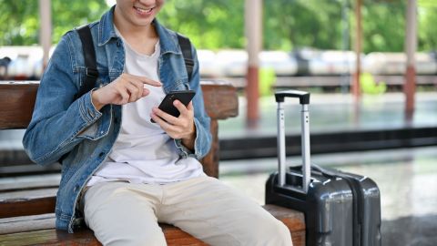 A photo of a person sitting on a bench using a smartphone with a suitcase next to them