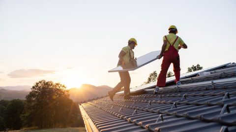 Two solar installers carry a solar panel to its new location on a roof.