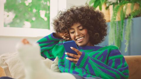 Young adult with a broken leg using a mobile phone in their apartment.