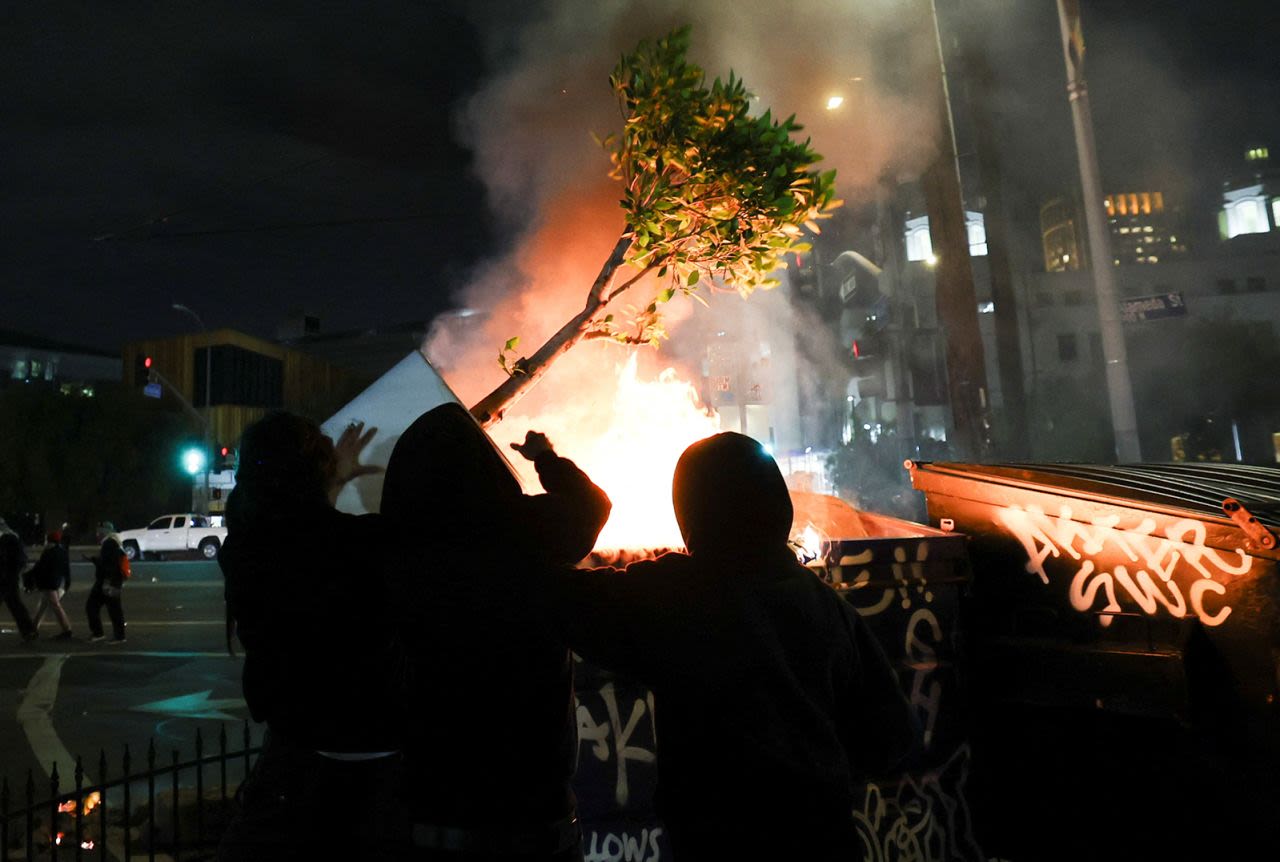 Los manifestantes arrojan una planta a un fuego que arde en un contenedor de basura durante una protesta contra las redadas federales de inmigración en el centro de Los Ángeles, California, el domingo