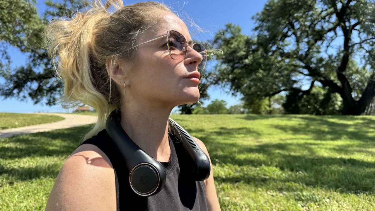 uma mulher loira e de óculos usa um ventilador de pescoço em um dia ensolarado em um parque
