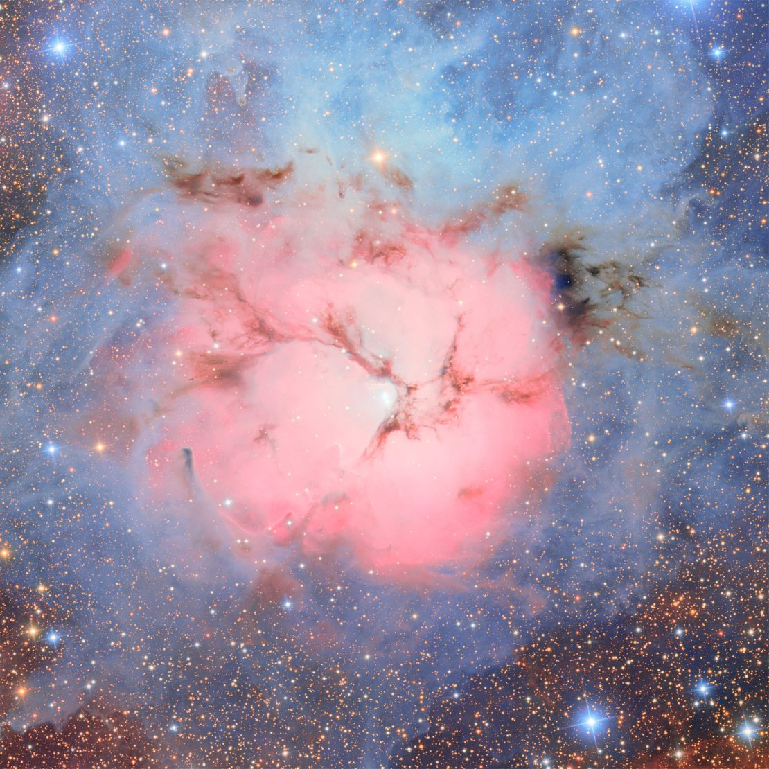 The Trifid Nebula is an unusual combination of an open cluster of stars, an emission nebula of glowing clouds of gas and dust that emits its own light (the pink region), a reflection nebula that reflects the light of nearby stars (the blue region), and a dark nebula so dense it blocks light from objects behind it (the dark regions).
