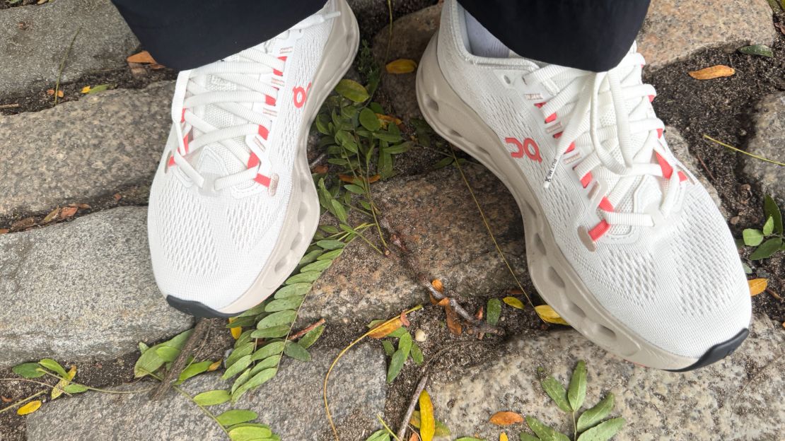 A person wearing white On Cloudsurfer shoes on a cobblestone road.