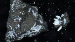 A microscope image of a dark Bennu particle, about a millimeter long, with a crust of bright phosphate. To the right is a smaller fragment that broke off.