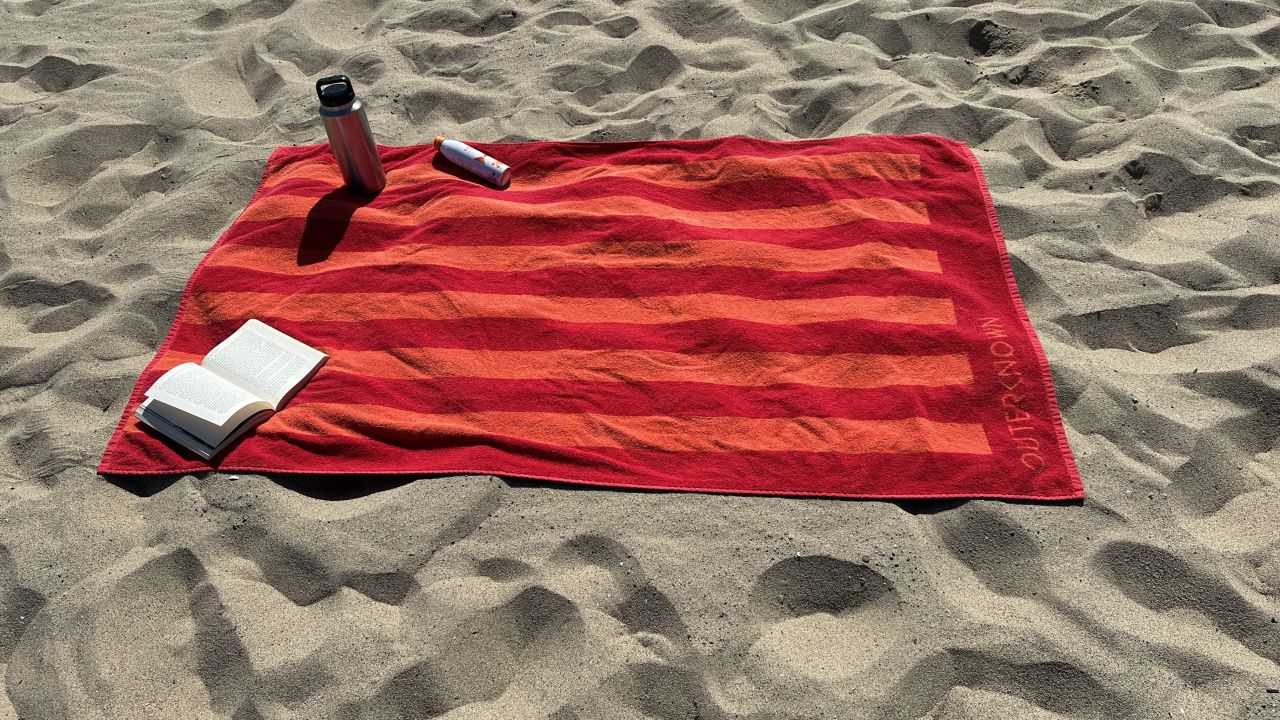 A photo of The Beach Towel from the brand Outerknown on a beach with various items on top of it