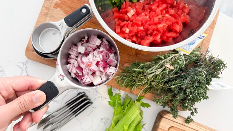 A hand holding up a measuring cup full of onion with other ingredients in the background