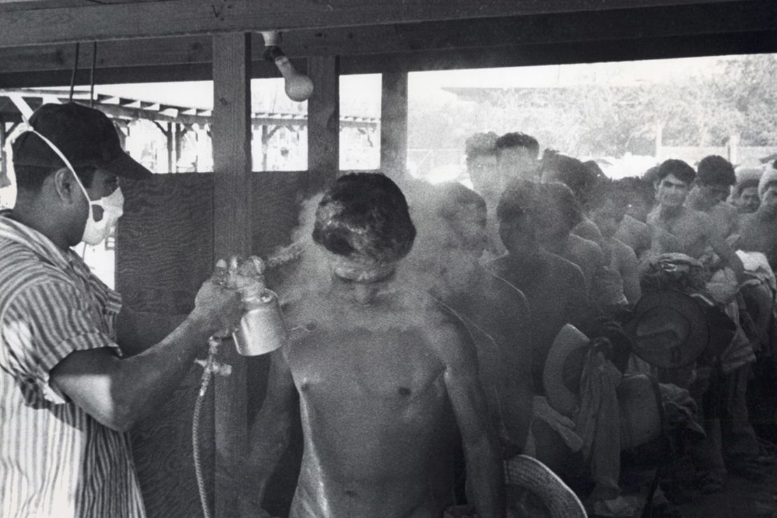 This photo taken in Hidalgo, Texas, in 1956 shows a masked worker spraying?<em>braceros</em>?with DDT while others wait in line.