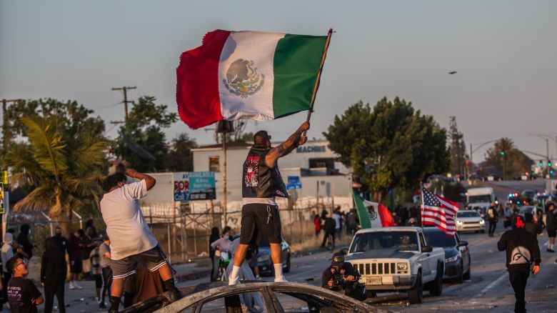 Un manifestante de pie sobre un auto incendiado, sosteniendo una bandera mexicana, en la Avenida Atlantic el 7 de junio de 2025 en Paramount, California.