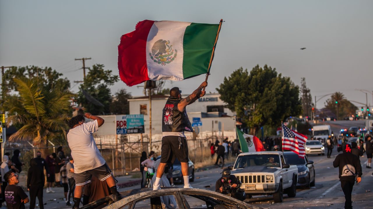 Un manifestante de pie sobre un auto incendiado, sosteniendo una bandera mexicana, en la Avenida Atlantic el 7 de junio de 2025 en Paramount, California.