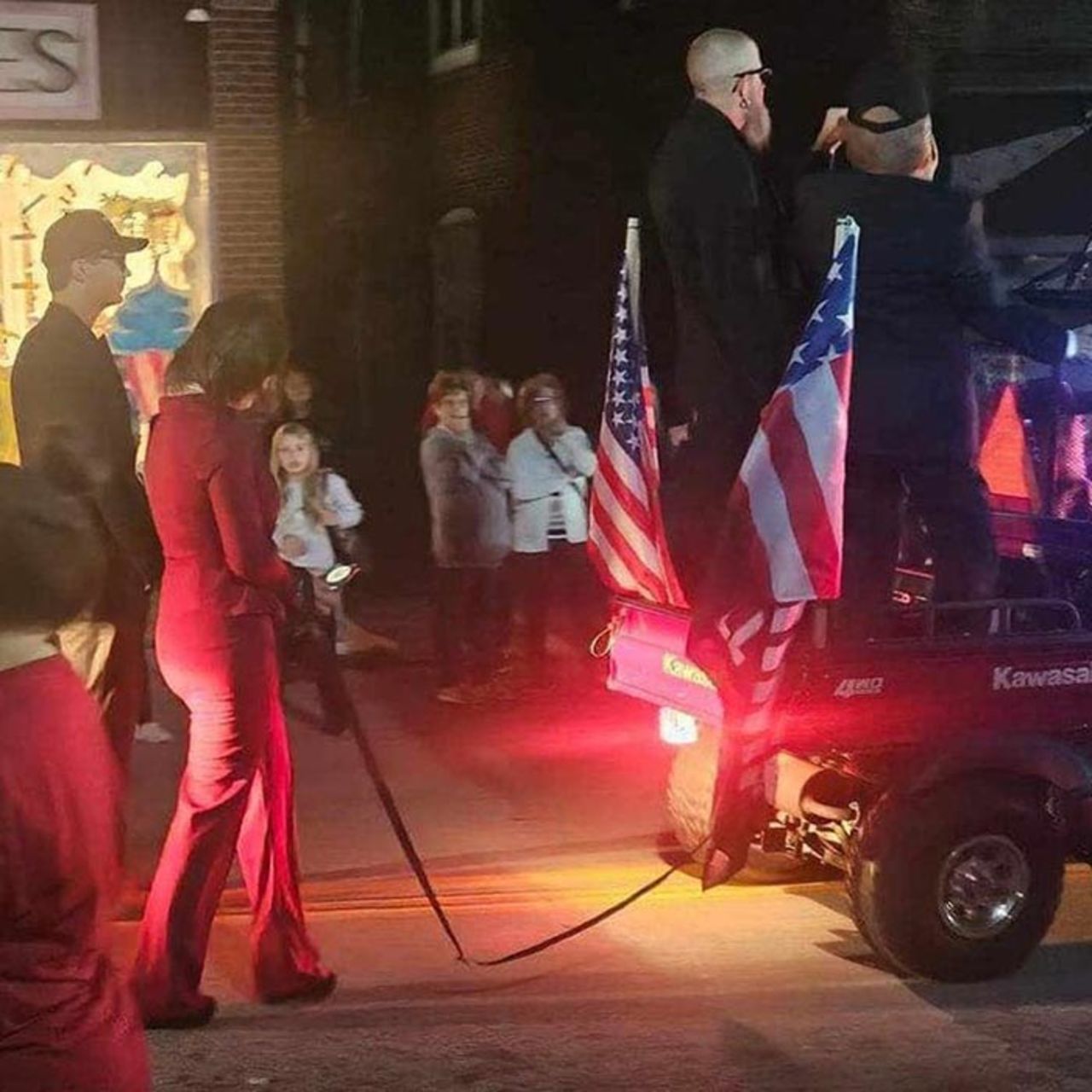 This photo shows a person wearing a red pantsuit — appearing to be dressed as Harris — walking with their hands bound and tied to the back of a golf cart flanked with American flags and a Trump campaign sign during a Halloween parade display in Pittsburgh.