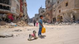 A Palestinian child in Gaza stands in the street with a water can and a toy horse, surrounded by rubble.