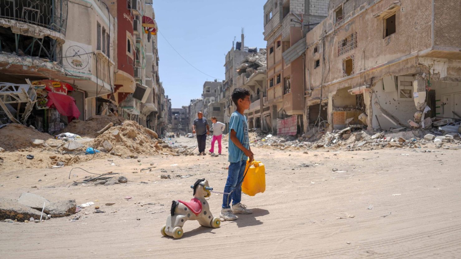 A Palestinian child stands in a rubble-strewn street in Gaza holding a yellow water jug in one hand and pulling a toy horse with the other.