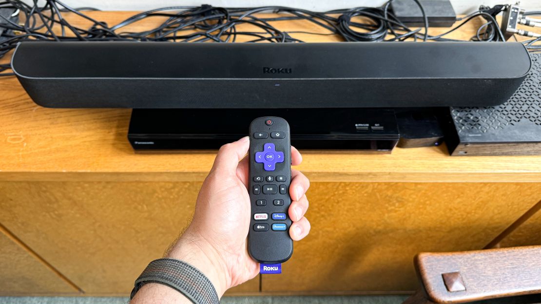 A hand holds a Roku remote in front of the Roku Streambar Pro.