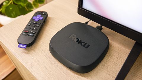 The Roku Ultra 2024 (right) and Roku Voice Remote Pro (left) sit on a countertop below a TV.