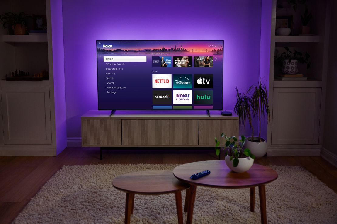 The Roku Ultra 2024 is connected to a TV on a countertop that's got the Roku home screen up, but at night with the TV glowing purple and tables in front of it.