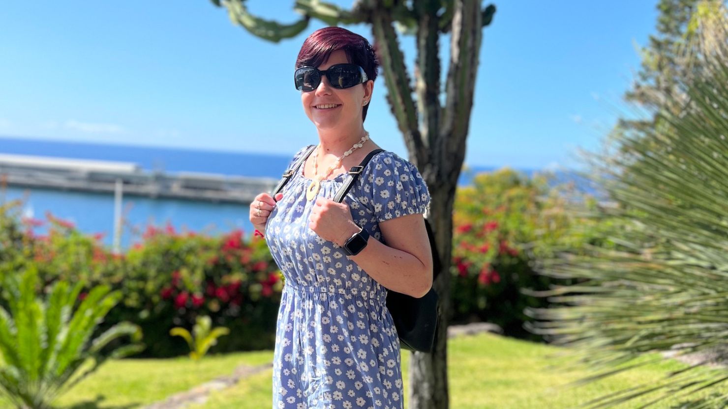 Sassy Wyatt is pictured smiling, holding a backpack, with blue sky, green grass and blue sea behind her.