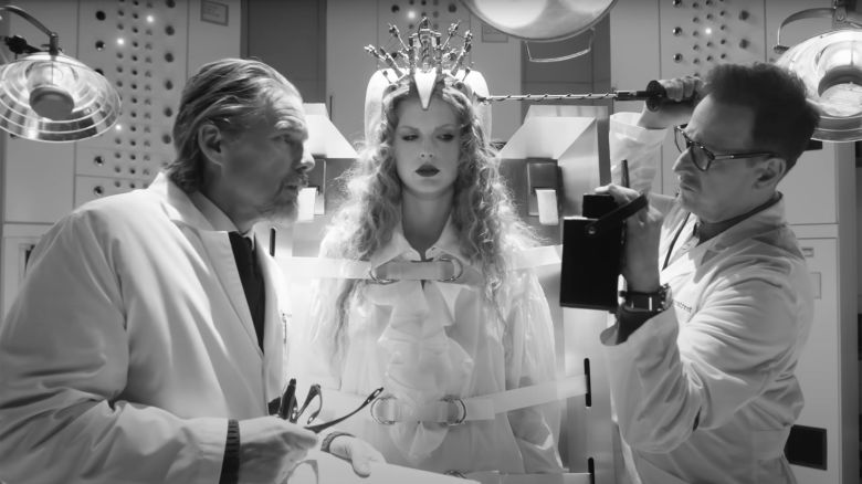 In a black and white screengrab, Ethan Hawke wears a white scientist's lab coat, while Taylor Swift appears strapped to a vertical gurney. 