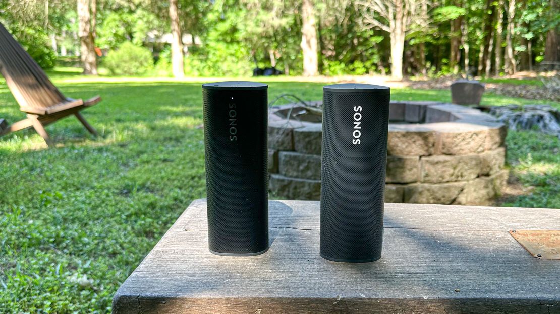 Two portable speakers, a Sonos Roam and a Sonos Roam 2, rest on a stone surface in a grassy backyard.