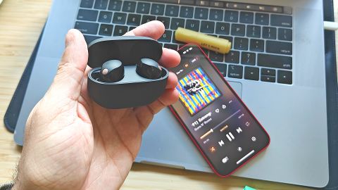 A hand holding encased wireless earbuds in front of a phone and laptop.