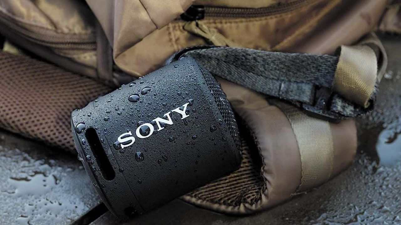 A small black speaker attached to a brown backpack rest on the ground in the rain.