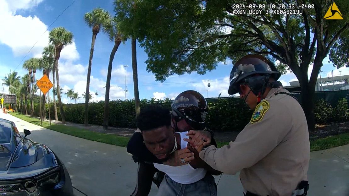 The officer puts his right arm around Hill and brings him down to sit on the sidewalk.