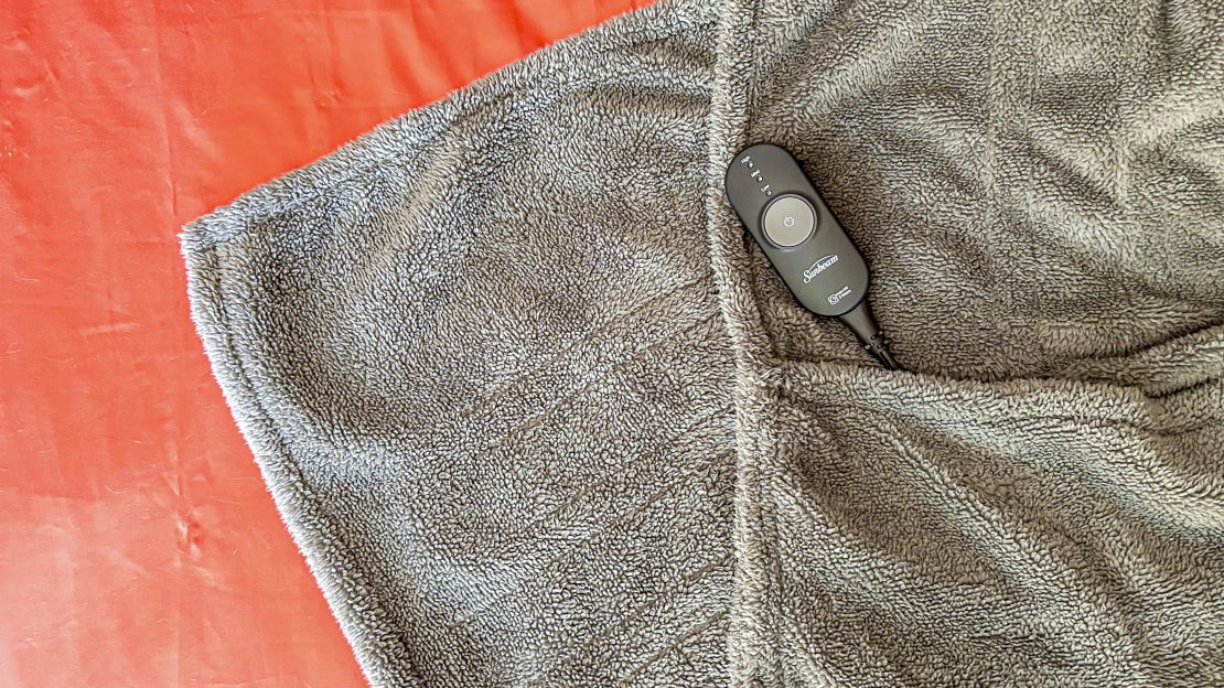 The Sunbeam Ultimate Lofttec Foot Pocket Electric Heated Throw and its remote flat on a bed.