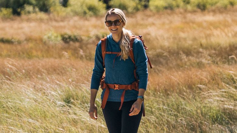 A person in a teal fleece hoodie with an orange backpack walks through a grassy area.