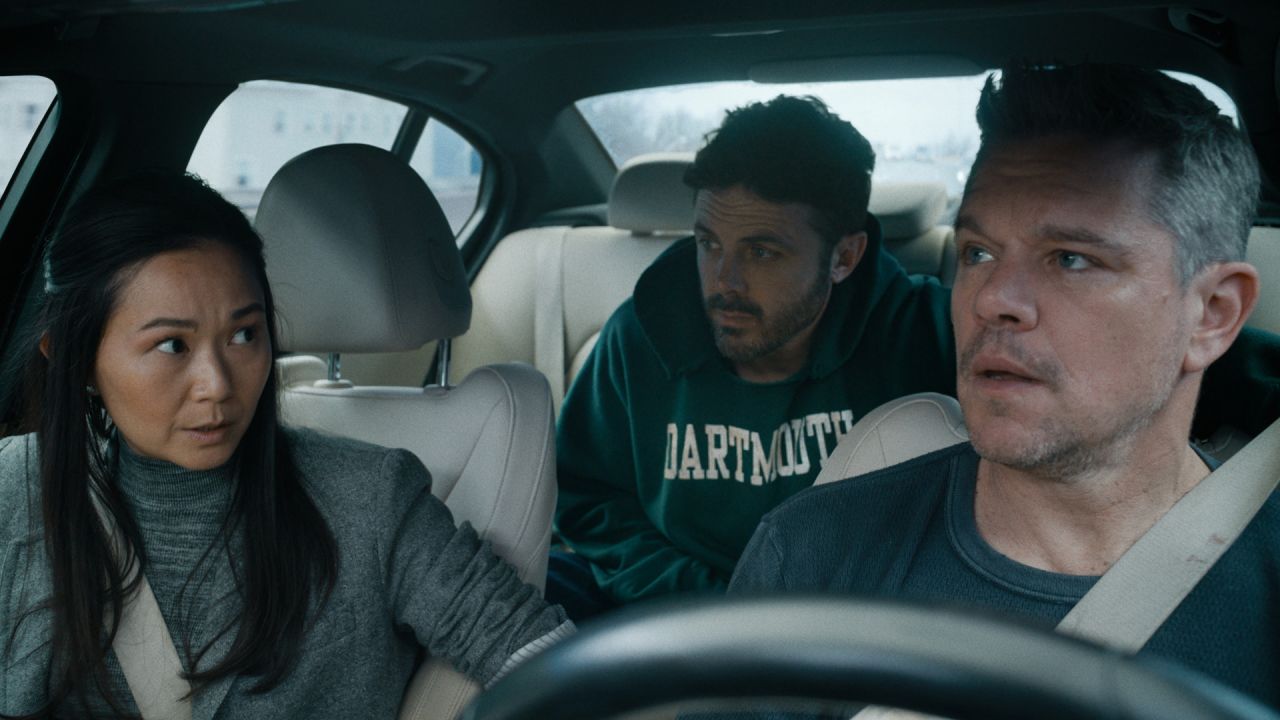 Matt Damon, Casey Affleck, and Hong Chau in a car in a scene from "The Instigators." The view is through the front windshield: Damon, at right, is behind the wheel and looking in the rear view mirror at Affleck in the back seat; Affleck is looking at Chau in the front passenger seat; Chau is looking at Damon. Chau and Damon are seatbelted; Affleck does not appear to have a seatbelt on.