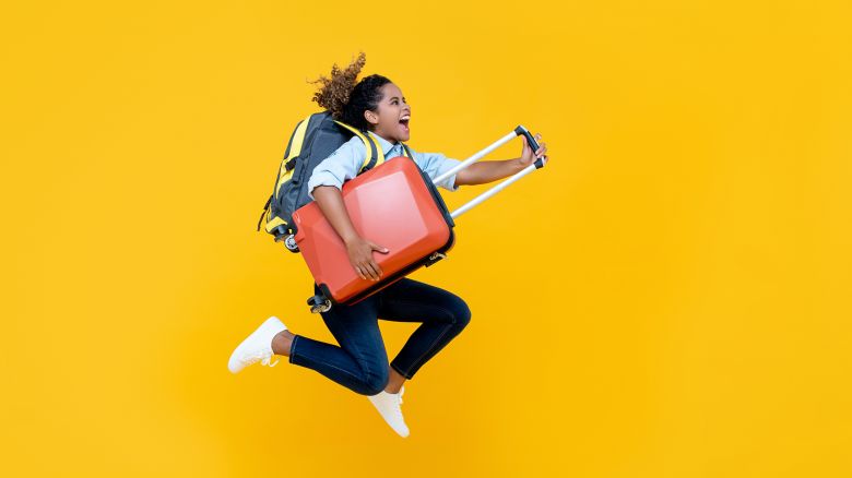 Excited African American woman tourist woman with backpack and luggage jumping in mid-air studio shot isolated on colorful yellow background