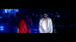 Usher, on stage wearing shades and a silver jumpsuit, sings as he's surrounded by male dancers.