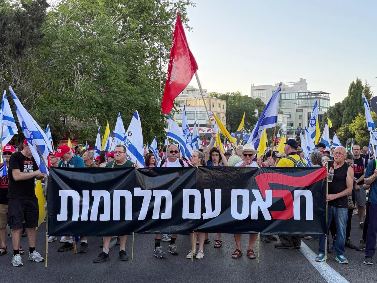 Protesters march along the streets of Haifa, Israel, carrying Israeli flags and yellow flags for the hostages. The sign says "Enough with the wars" following Israel's 12-day conflict with Iran and the war in Gaza, which has surpassed 630 days. The red flag is an anti-government protest symbol.