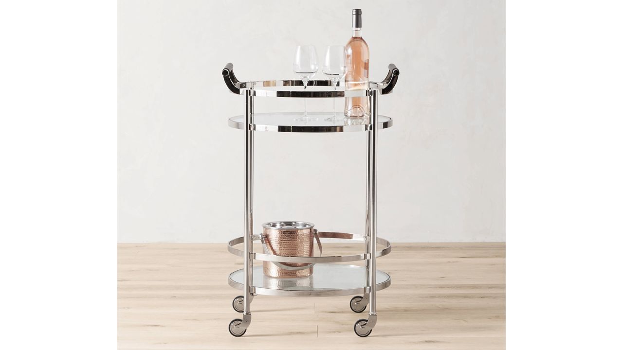 The Williams Sonoma Truman Round Bar Cart with a bottle of wine and two glasses on its top shelf and an ice bucket on its bottom shelf.