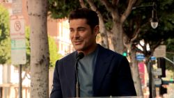 Zac Efron stands at the podium mic at his Hollywood Walk of Fame ceremony. He's wearing a navy blue blazer over a silky gray t-shirt, and smiling slightly as he addresses the crowd.